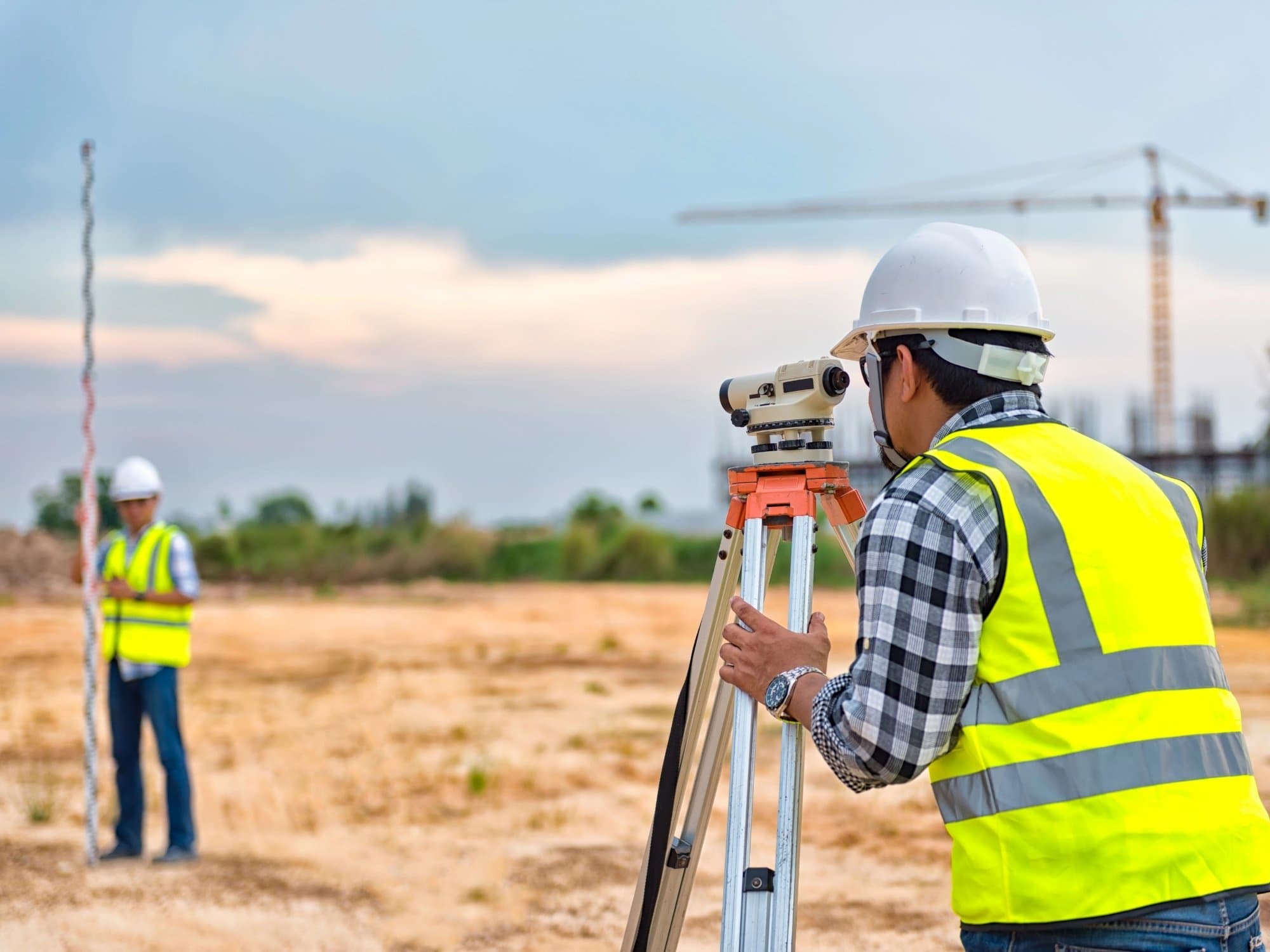 A surveyor taking measurements through a level on a tripod while a colleague holds the staff